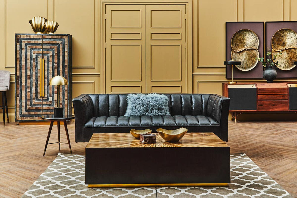 A stylish black leather three-seater sofa with flared arms is positioned in a cozy living room setting. The sofa features a tufted design and is complemented by a light blue throw pillow. In front of the sofa, a dark wooden coffee table holds decorative bowls. The background includes a textured wall with a large piece of artwork and an ornate cabinet, adding to the room's elegant ambiance. A patterned