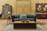 A stylish black leather three-seater sofa with flared arms is positioned in a cozy living room setting. The sofa features a tufted design and is complemented by a light blue throw pillow. In front of the sofa, a dark wooden coffee table holds decorative bowls. The background includes a textured wall with a large piece of artwork and an ornate cabinet, adding to the room's elegant ambiance. A patterned