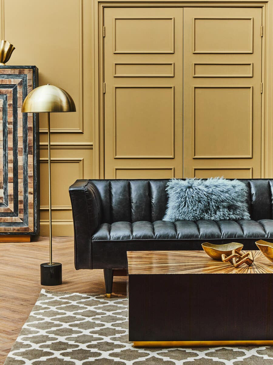 A stylish black leather three-seater sofa with flared arms, positioned in a cozy living space. The sofa features a tufted design and is adorned with a light blue throw blanket. A wooden coffee table sits in front, decorated with a few items. The background includes a patterned rug and a tall floor lamp, with a decorative wall panel behind the sofa