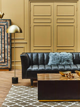 A stylish black leather three-seater sofa with flared arms, positioned in a cozy living space. The sofa features a tufted design and is adorned with a light blue throw blanket. A wooden coffee table sits in front, decorated with a few items. The background includes a patterned rug and a tall floor lamp, with a decorative wall panel behind the sofa