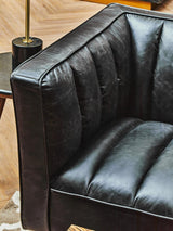 Black leather sofa featuring flared arms and a tufted design on the seat and back cushions, set against a wooden floor with a side table visible in the background