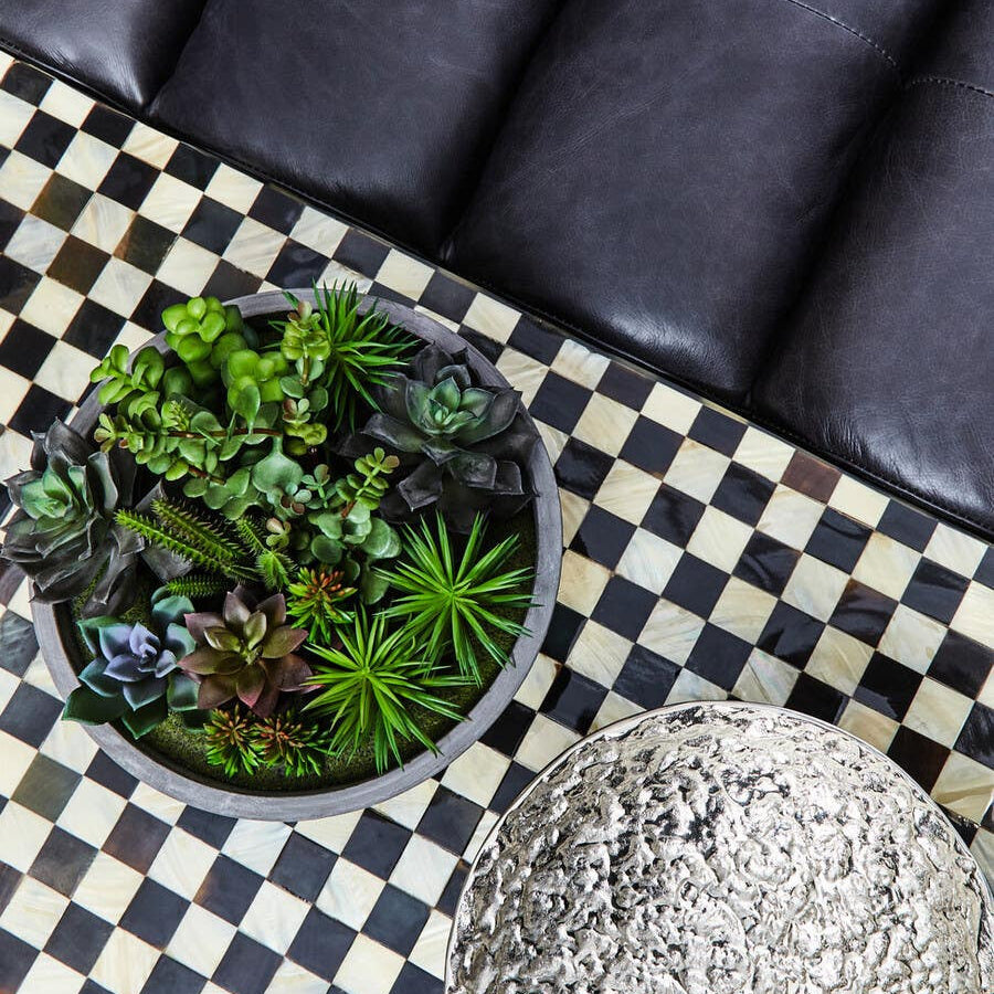Antique black leather modular sofa chair with a smooth, armless design, positioned next to a round planter filled with green plants, on a black and white checkered rug
