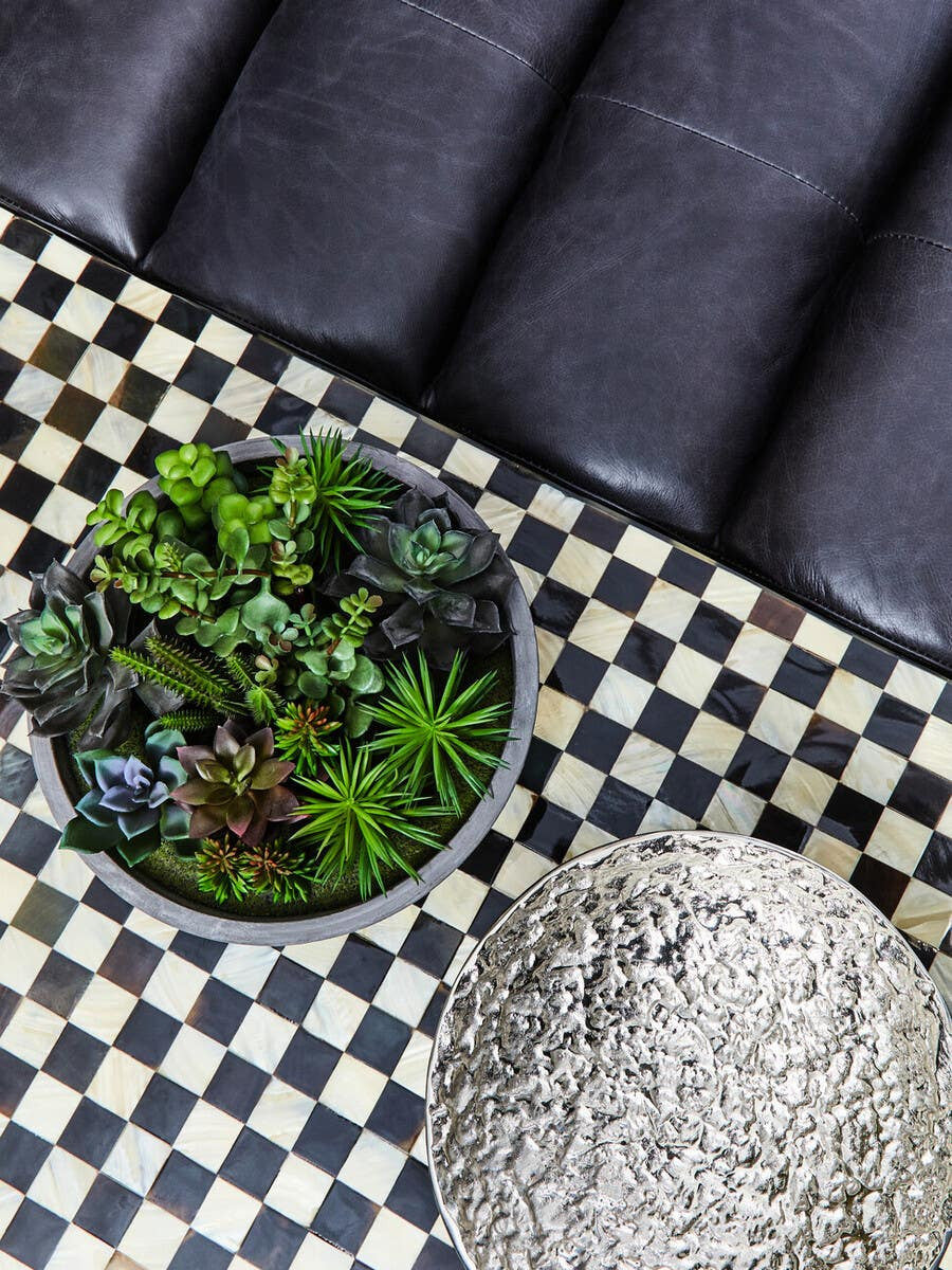 Antique black leather modular sofa chair with a smooth, armless design, positioned next to a round planter filled with green plants, on a black and white checkered rug