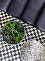 Antique black leather modular sofa chair with a smooth, armless design, positioned next to a round planter filled with green plants, on a black and white checkered rug