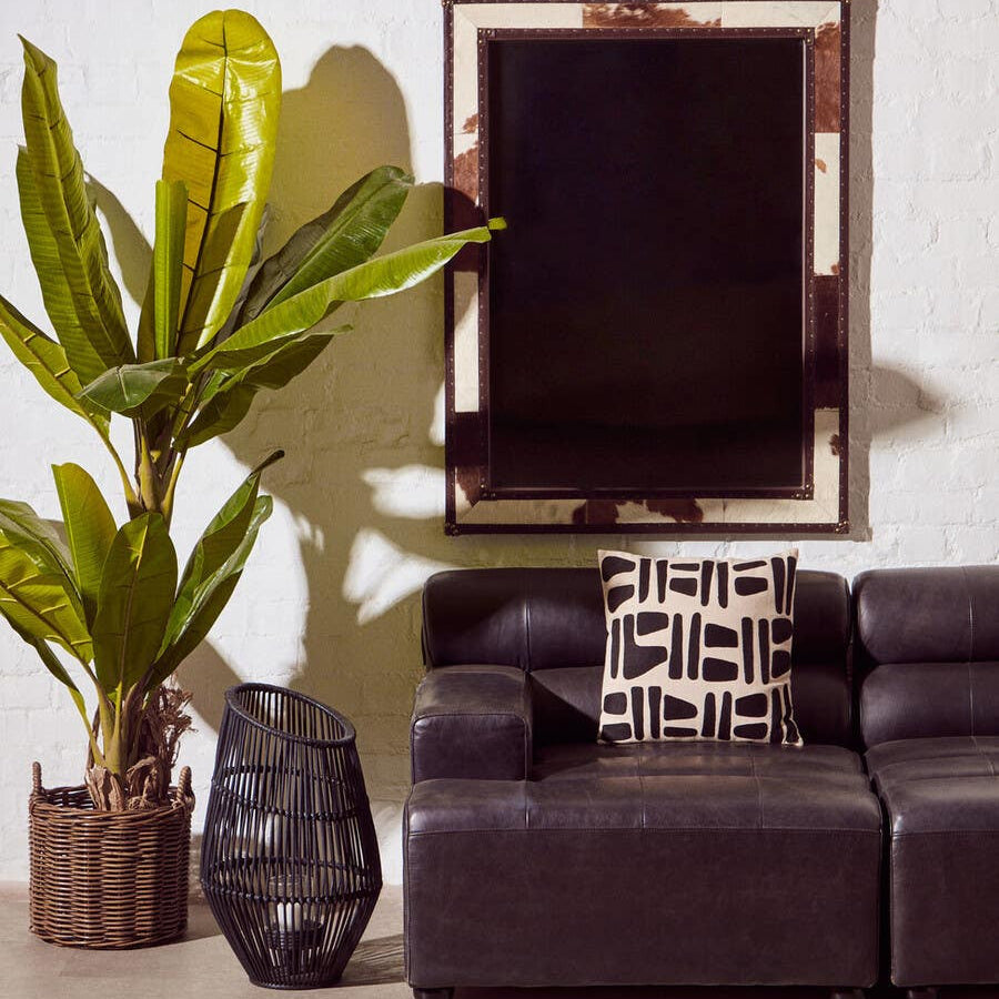 Left arm leather modular chaise sofa in antique black, featuring a sleek design with plush cushions. Positioned next to a tall green plant in a woven basket and a decorative black vase. A large mirror hangs on the wall behind the sofa, reflecting the room's light. The floor is covered with a textured area rug