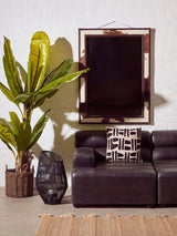 Left arm leather modular chaise sofa in antique black, featuring a sleek design with plush cushions. Positioned next to a tall green plant in a woven basket and a decorative black vase. A large mirror hangs on the wall behind the sofa, reflecting the room's light. The floor is covered with a textured area rug