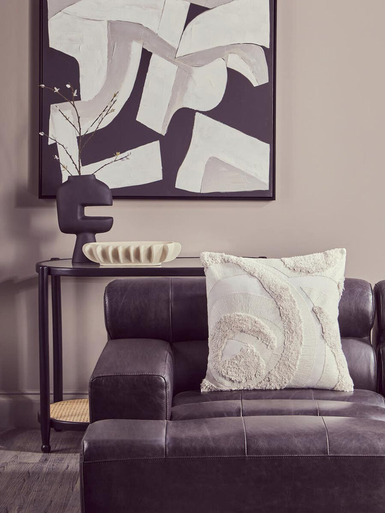 Antique black left arm leather modular chaise sofa with a decorative white pillow, positioned next to a sleek side table and a modern lamp, against a neutral wall featuring abstract artwork