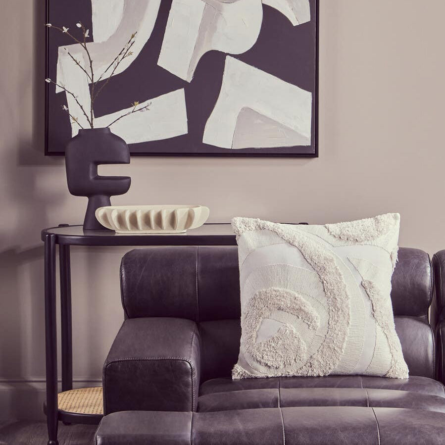 Antique black left arm leather modular chaise sofa with a decorative white pillow, positioned next to a sleek side table and a modern lamp, against a neutral wall featuring abstract artwork