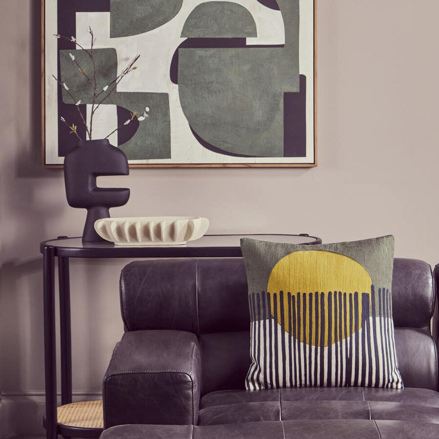 Antique black left arm leather modular chaise sofa with a textured surface, accompanied by a decorative pillow featuring a yellow sun design. A modern lamp and a console table with decorative items are visible in the background, along with an abstract wall art piece in muted colors