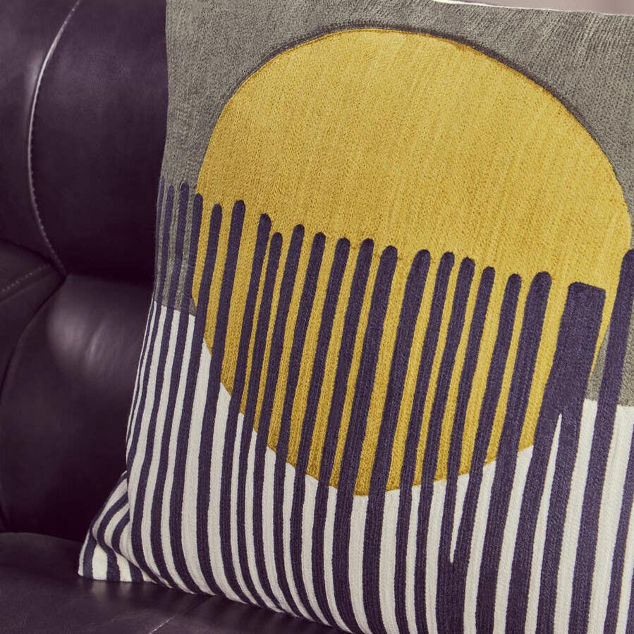 A close-up view of a decorative pillow featuring a yellow circular design with vertical stripes in shades of blue and white, placed on a dark leather chaise sofa. The sofa has a sleek, modern design with a left armrest, showcasing its antique black finish