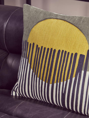 A close-up view of a decorative pillow featuring a yellow circular design with vertical stripes in shades of blue and white, placed on a dark leather chaise sofa. The sofa has a sleek, modern design with a left armrest, showcasing its antique black finish