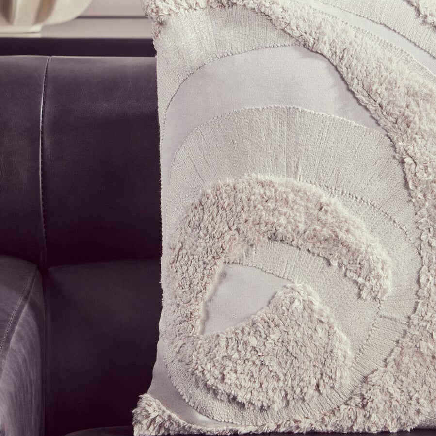A close-up view of a textured cream-colored throw pillow with a spiral design, resting on a dark leather chaise sofa. The sofa features a sleek, modern design with a left arm, showcasing the rich antique black leather upholstery. The background is softly blurred, emphasizing the sofa and pillow