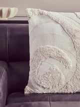 A close-up view of a textured cream-colored throw pillow with a spiral design, resting on a dark leather chaise sofa. The sofa features a sleek, modern design with a left arm, showcasing the rich antique black leather upholstery. The background is softly blurred, emphasizing the sofa and pillow