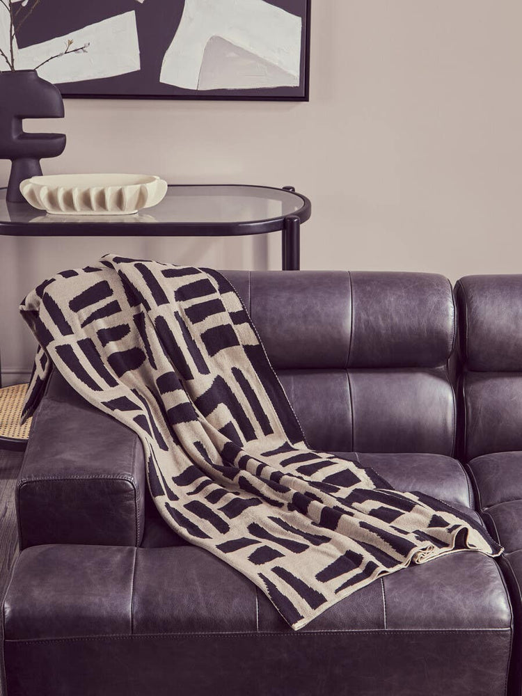 Antique black leather modular chaise sofa with a left arm, featuring a modern geometric patterned throw blanket draped over the seat. The sofa has a sleek design with plush cushions and a contemporary aesthetic, set against a neutral wall with abstract artwork in the background