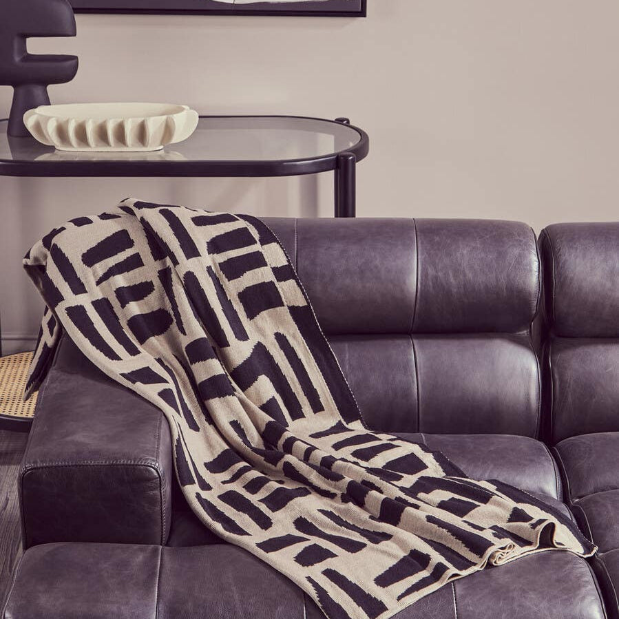 Antique black leather modular chaise sofa with a left arm, featuring a modern geometric patterned throw blanket draped over the seat. The sofa has a sleek design with plush cushions and a contemporary aesthetic, set against a neutral wall with abstract artwork in the background
