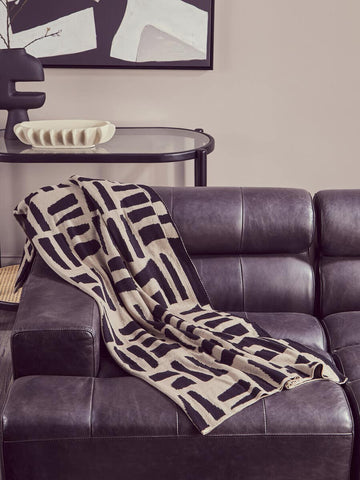 Antique black leather modular chaise sofa with a left arm, featuring a modern geometric patterned throw blanket draped over the seat. The sofa has a sleek design with plush cushions and a contemporary aesthetic, set against a neutral wall with abstract artwork in the background