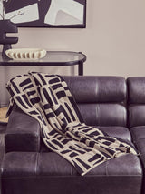 Antique black leather modular chaise sofa with a left arm, featuring a modern geometric patterned throw blanket draped over the seat. The sofa has a sleek design with plush cushions and a contemporary aesthetic, set against a neutral wall with abstract artwork in the background