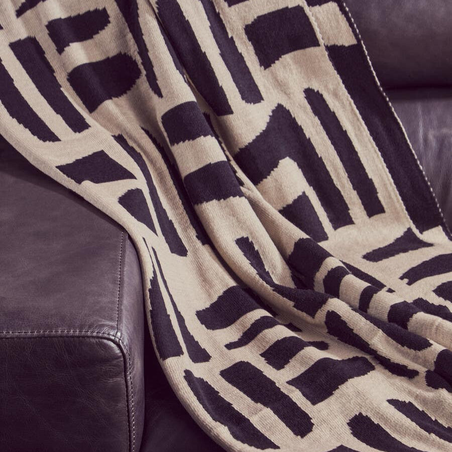 A close-up view of a patterned throw blanket draped over a dark leather sofa, featuring geometric designs in black and beige. The texture of the leather is visible, highlighting the luxurious feel of the chaise