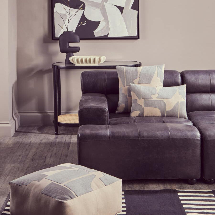 Antique black left arm leather modular chaise sofa with a contemporary design, featuring plush cushions and a sleek silhouette. A decorative throw pillow rests on the sofa, and a geometric-patterned pouf is placed nearby on a textured area rug. A modern artwork hangs on the wall in the background, complementing the overall aesthetic