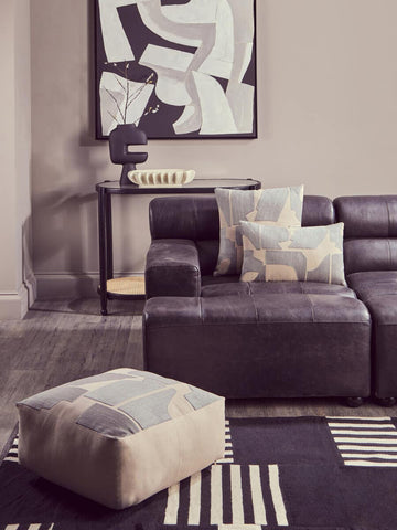 Antique black left arm leather modular chaise sofa with a contemporary design, featuring plush cushions and a sleek silhouette. A decorative throw pillow rests on the sofa, and a geometric-patterned pouf is placed nearby on a textured area rug. A modern artwork hangs on the wall in the background, complementing the overall aesthetic