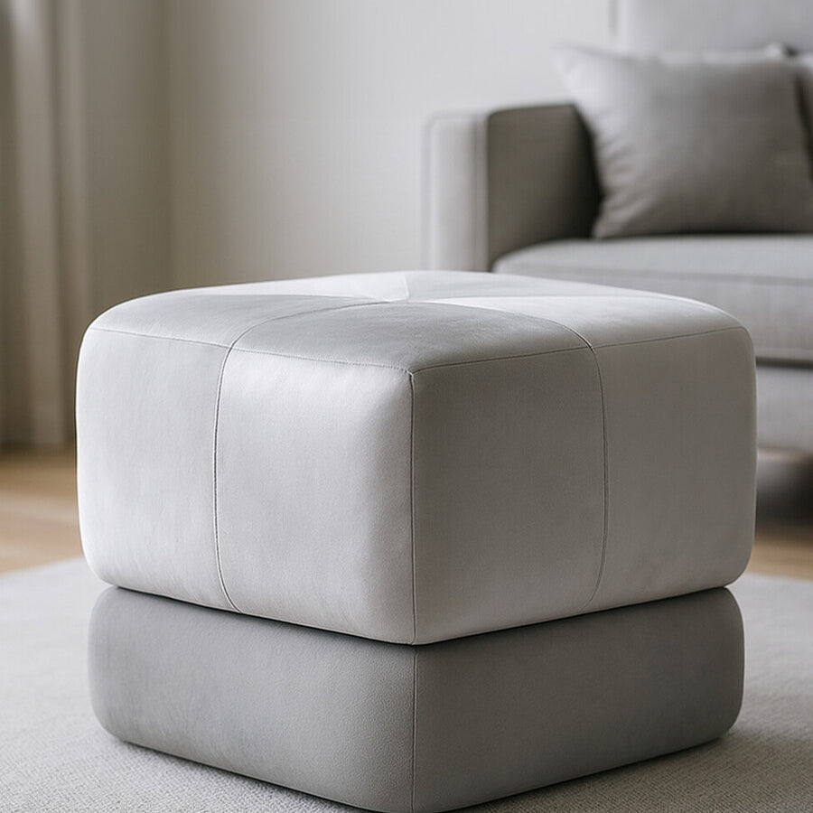 Square stool upholstered in grey velvet, featuring a two-tone design with a lighter top and darker base. The stool is placed on a light-colored rug in a bright room with a sofa in the background. Natural light illuminates the space, highlighting the soft texture of the fabric