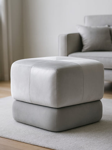 Square stool upholstered in grey velvet, featuring a two-tone design with a lighter top and darker base. The stool is placed on a light-colored rug in a bright room with a sofa in the background. Natural light illuminates the space, highlighting the soft texture of the fabric