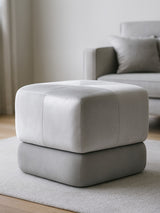 Square stool upholstered in grey velvet, featuring a two-tone design with a lighter top and darker base. The stool is placed on a light-colored rug in a bright room with a sofa in the background. Natural light illuminates the space, highlighting the soft texture of the fabric