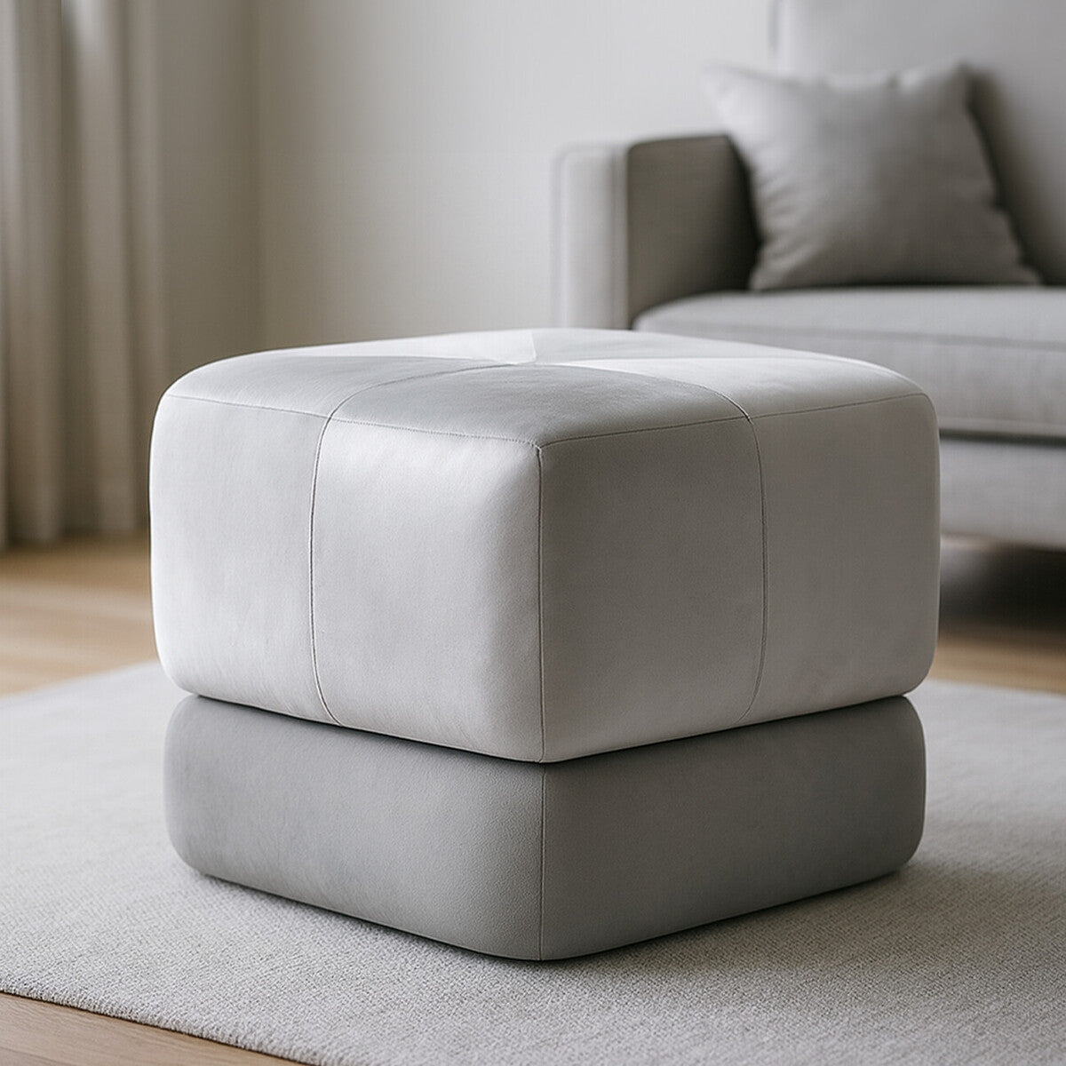 Square stool upholstered in grey velvet, featuring a two-tone design with a lighter top and darker base, placed on a light-colored rug in a modern living space with a soft sofa in the background