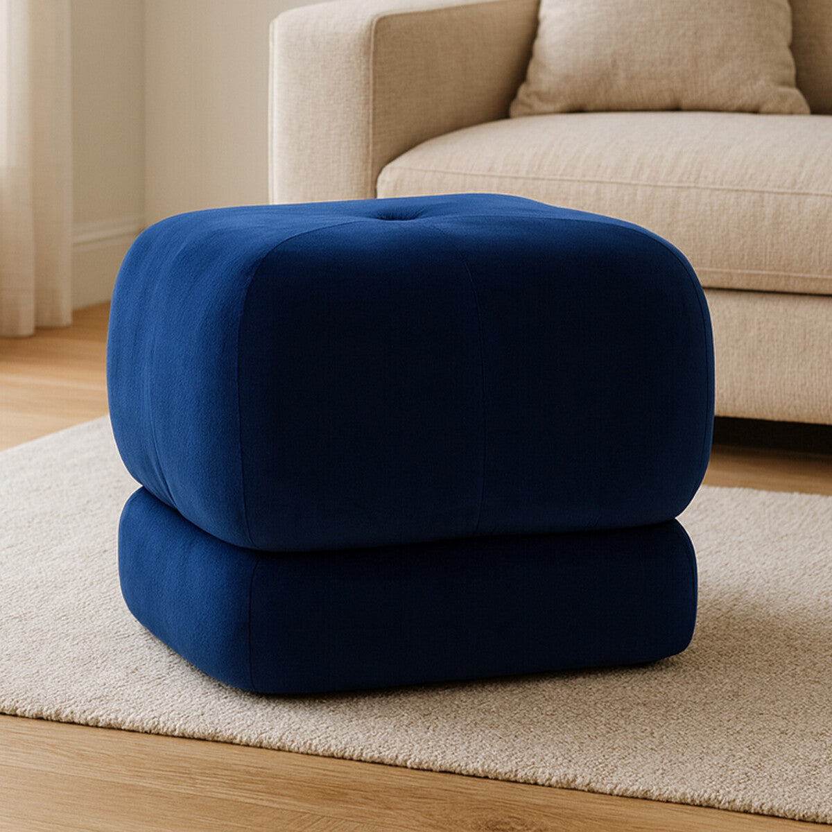 Midnight blue velvet square stool with a soft, plush texture and a rounded top, featuring a button detail in the center, placed on a light wooden floor in a cozy living room setting. A beige sofa is visible in the background