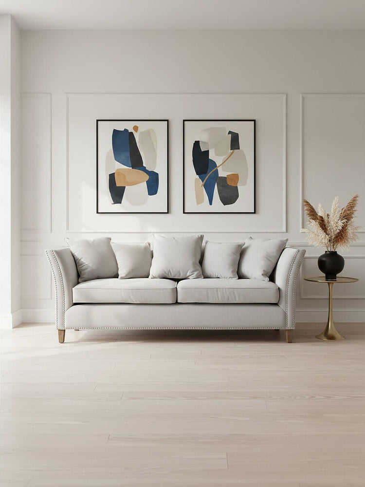 Three-seat grey fabric sofa with plush cushions, positioned against a light-colored wall. Above the sofa are two framed abstract art pieces featuring blue and beige designs. A decorative plant in a vase is placed on a nearby side table, complementing the modern aesthetic of the room. The flooring is light wood, enhancing the overall bright and airy atmosphere