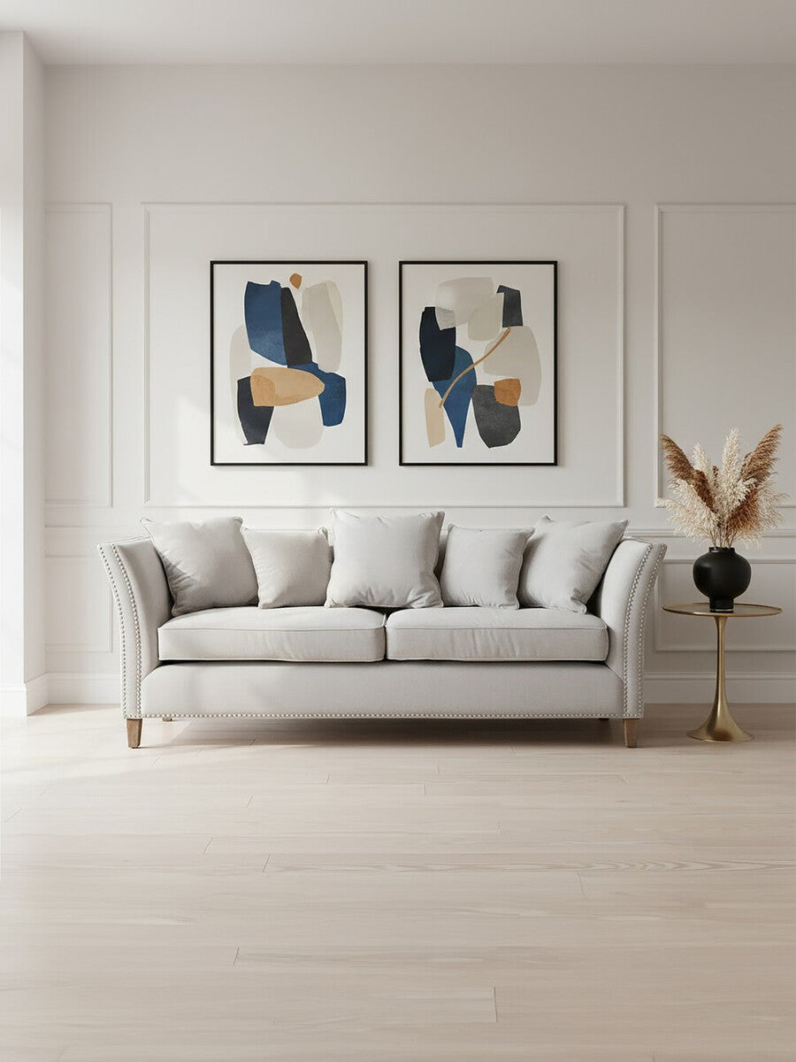 Three-seat grey fabric sofa with plush cushions, positioned against a light-colored wall. Above the sofa are two framed abstract art pieces featuring blue and beige designs. A decorative plant in a vase is placed on a nearby side table, complementing the modern aesthetic of the room. The flooring is light wood, enhancing the overall bright and airy atmosphere
