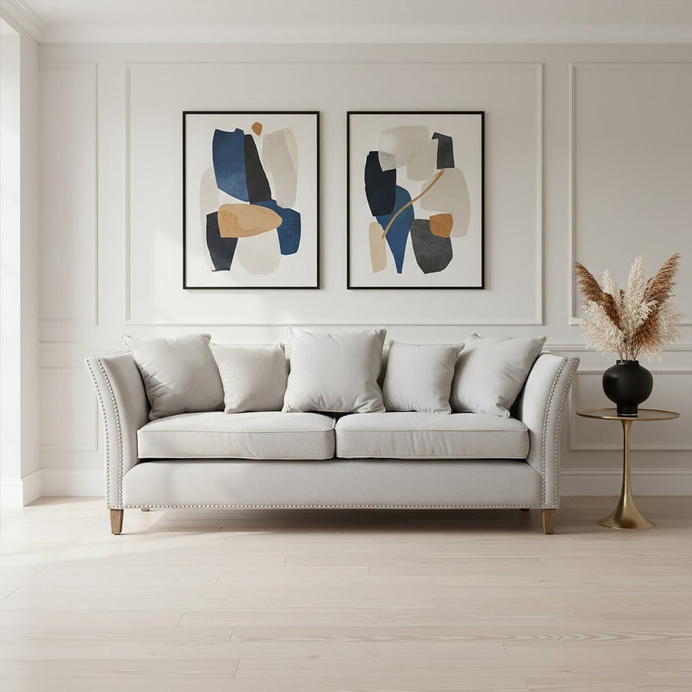 Three-seat grey fabric sofa with plush cushions, positioned against a light-colored wall. Two abstract art pieces in black and blue frames are displayed above the sofa. A decorative plant in a black vase stands to the right of the sofa, and the floor features light wood planks