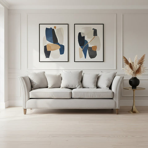 Three-seat grey fabric sofa with plush cushions, positioned against a light-colored wall. Two abstract art pieces in black and blue frames are displayed above the sofa. A decorative plant in a black vase stands to the right of the sofa, and the floor features light wood planks