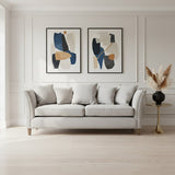 Three-seat grey fabric sofa with plush cushions, positioned against a light-colored wall. Two abstract art pieces in black and blue frames are displayed above the sofa. A decorative plant in a black vase stands to the right of the sofa, and the floor features light wood planks