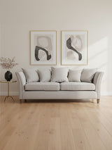 Three-seat grey fabric sofa with plush cushions and a modern design, positioned against a light-colored wall. Two abstract framed artworks hang above the sofa, and a small decorative plant in a vase is placed next to a black floor lamp on the left. The flooring is light wood, creating a warm and inviting atmosphere