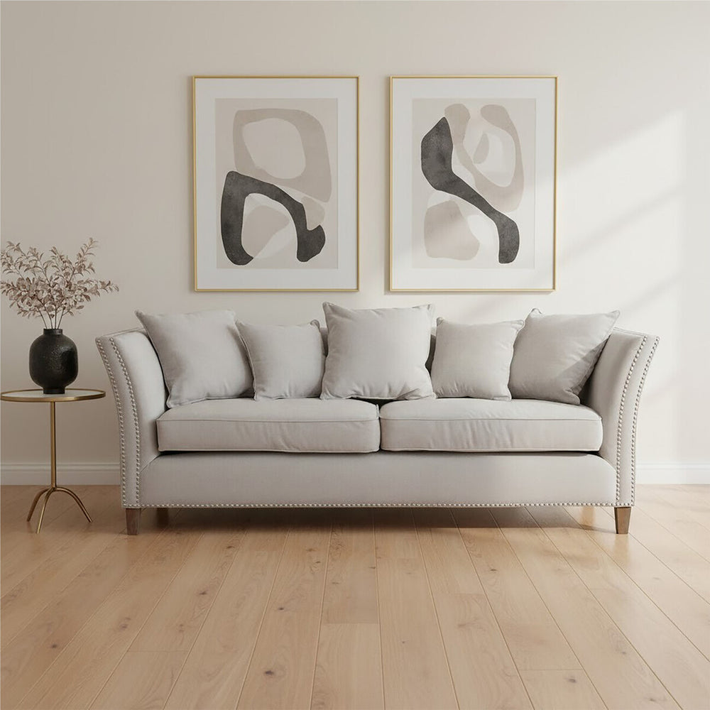 Three-seat grey fabric sofa with plush cushions, featuring a classic design and wooden legs, positioned in a bright room with light wooden flooring. Two abstract art pieces in gold frames hang on the wall behind the sofa, and a small black side table with a decorative vase is placed beside it