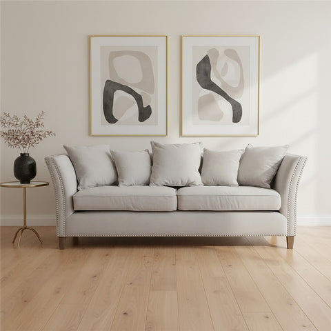 Three-seat grey fabric sofa with plush cushions, featuring a classic design and wooden legs, positioned in a bright room with light wooden flooring. Two abstract art pieces in gold frames hang on the wall behind the sofa, and a small black side table with a decorative vase is placed beside it