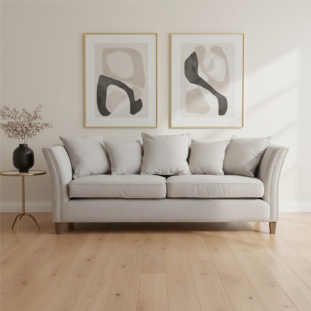 Three-seat grey fabric sofa with plush cushions, featuring a classic design and wooden legs, positioned in a bright room with light wooden flooring. Two abstract art pieces in gold frames hang on the wall behind the sofa, and a small black side table with a decorative vase is placed beside it