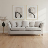 Three-seat grey fabric sofa with plush cushions, featuring a classic design and wooden legs, positioned in a bright room with light wooden flooring. Two abstract art pieces in gold frames hang on the wall behind the sofa, and a small black side table with a decorative vase is placed beside it