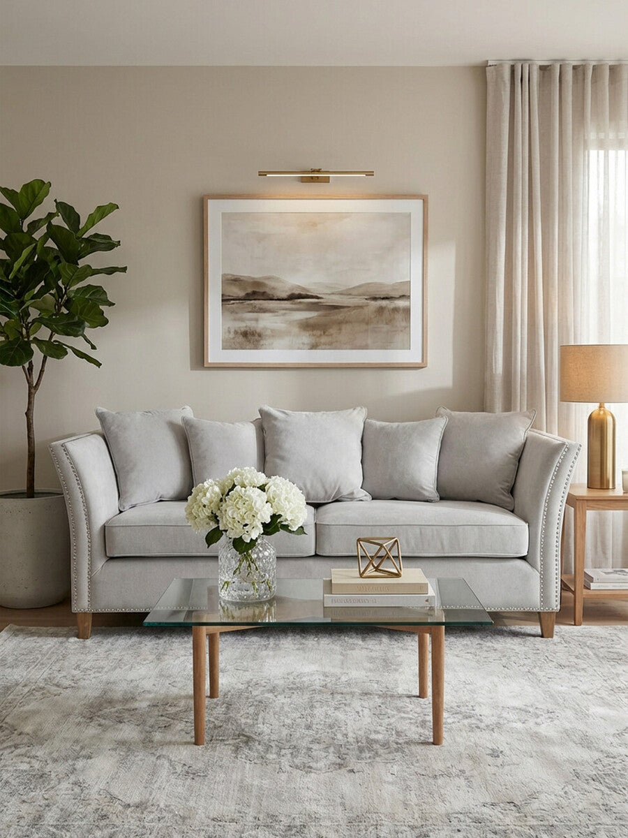Three-seat grey fabric sofa with plush cushions, positioned in a cozy living room setting. A wooden coffee table with a decorative book and a vase of white flowers sits in front of the sofa. Soft, neutral-colored walls and a large piece of artwork are visible in the background, along with a floor lamp and a potted plant. Natural light filters in through a window with sheer curtains