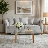 Three-seat grey fabric sofa with plush cushions, positioned in a cozy living room setting. A glass coffee table with a decorative book and a vase of white flowers sits in front of the sofa. Soft, neutral-toned area rug beneath the furniture, with a potted plant in the corner and light-colored curtains framing a window. A piece of abstract wall art hangs above the sofa, adding a modern touch