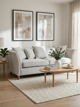 Three-seat grey fabric sofa with plush cushions, positioned in a bright living room. The sofa is complemented by a light wooden coffee table and decorative elements, including a potted plant and framed artwork on the walls. Natural light filters through sheer curtains, enhancing the cozy atmosphere. A soft area rug is placed beneath the furniture