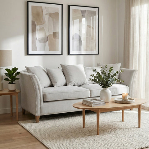 Three-seat grey fabric sofa with plush cushions, positioned in a bright living room. The sofa is accompanied by a light wooden coffee table and a decorative plant. Two framed artworks hang on the wall behind the sofa, and natural light filters through sheer curtains. A soft area rug is placed under the furniture, enhancing the cozy atmosphere