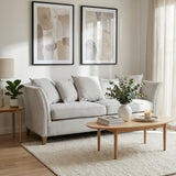 Three-seat grey fabric sofa with plush cushions, positioned in a bright living room. The sofa is accompanied by a light wooden coffee table and a decorative plant. Two framed artworks hang on the wall behind the sofa, and natural light filters through sheer curtains. A soft area rug is placed under the furniture, enhancing the cozy atmosphere