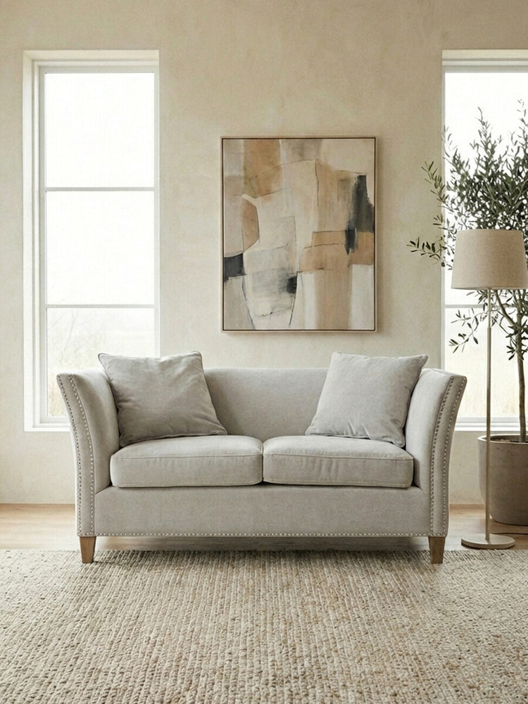 Two-seat sofa upholstered in light grey fabric, featuring two plush back cushions and two seat cushions. The sofa has rounded arms and wooden legs. In the background, there are two large windows allowing natural light, a piece of abstract wall art, and a potted plant beside the sofa, all set on a textured area rug