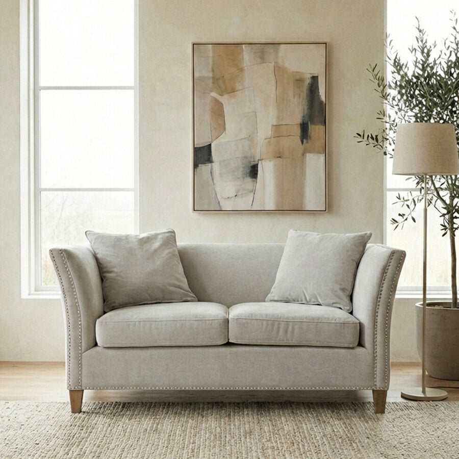 Two-seat sofa upholstered in light grey fabric, featuring two plush back cushions and two seat cushions. The sofa has rounded arms and wooden legs. In the background, there are two large windows allowing natural light, a piece of abstract wall art, and a potted plant beside the sofa, all set on a textured area rug