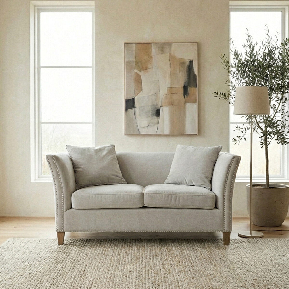 Two-seat grey fabric sofa with plush cushions, featuring a classic design and curved armrests, positioned in a bright room with large windows. A piece of abstract artwork hangs on the wall behind the sofa, and a tall floor lamp and potted plant are visible to the side, enhancing the cozy atmosphere. The sofa is placed on a light-colored rug