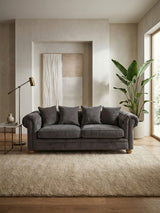 Three-seat grey fabric sofa with plush cushions, set against a light-colored wall and large windows that allow natural light to fill the room. A floor lamp and a potted plant are visible in the background, with a textured area rug beneath the sofa