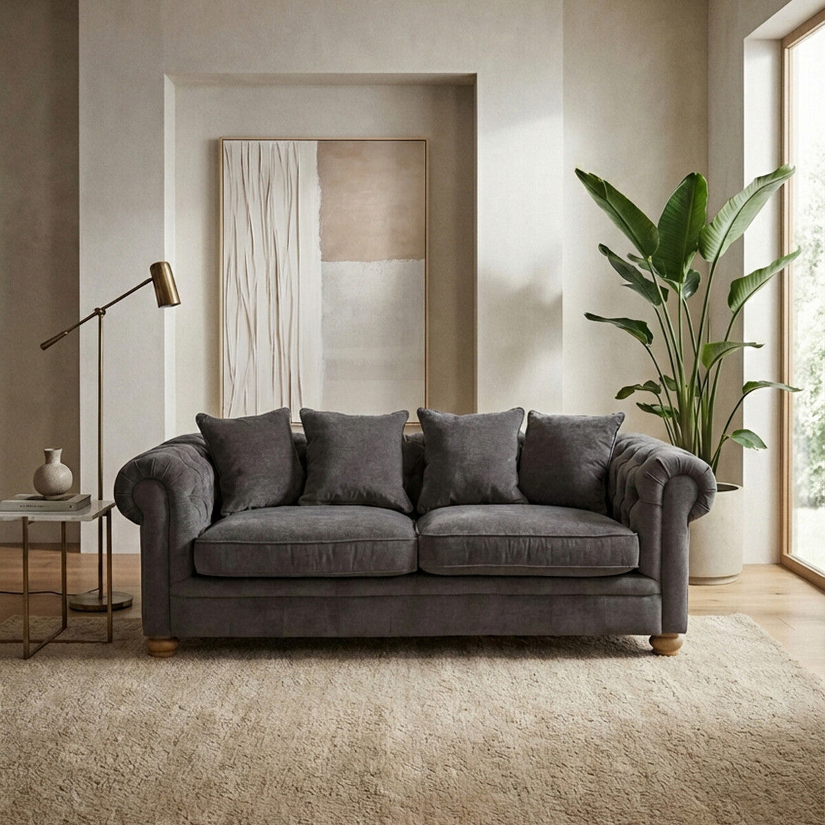 Three-seat grey fabric sofa with plush cushions, set against a light-colored wall and a large window. A tall indoor plant is visible to the right, and a floor lamp is positioned to the left. The sofa features a classic design with rolled arms and wooden legs, placed on a textured area rug