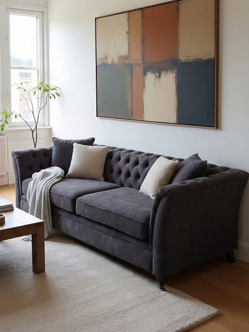 Three-seat grey velvet sofa with tufted backrest and armrests, adorned with two decorative pillows, placed in a bright room featuring a large abstract wall art piece and a wooden coffee table on a light rug